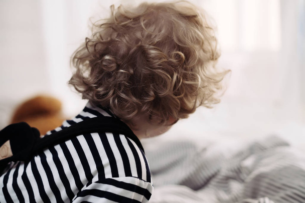 curly blonde little baby boy sitting on the bed playing - Fotó, kép