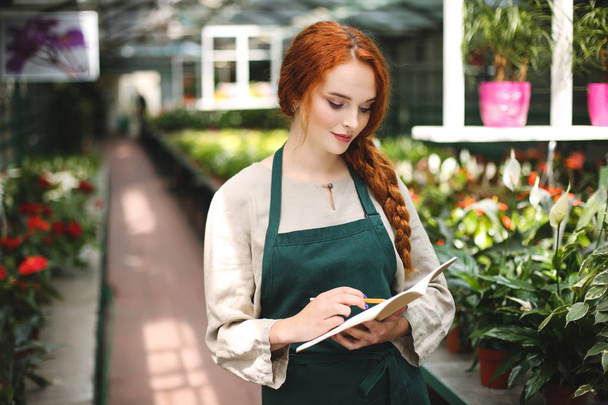Beautiful florist in apron standing with pencil in hand and dreamily writing in notepad while working in greenhouse - Photo, Image