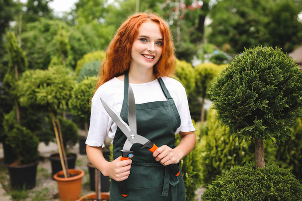 Красивая женщина с рыжими вьющимися волосами, стоящая в фартуке и держа большие садовые ножницы, счастливо глядя в камеру с растениями на заднем плане
 - Фото, изображение