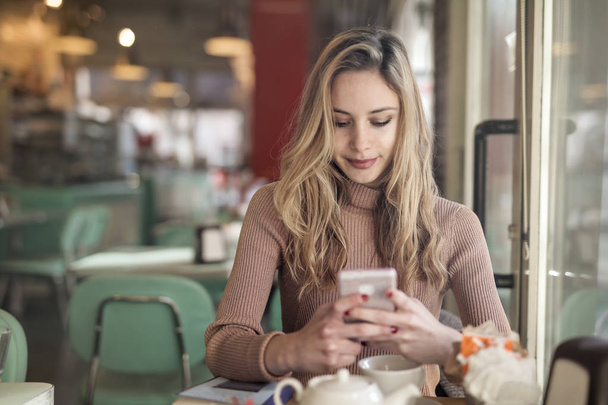 Young blonde woman sitting at a table and checking her smartphone in a bistro. - Photo, Image