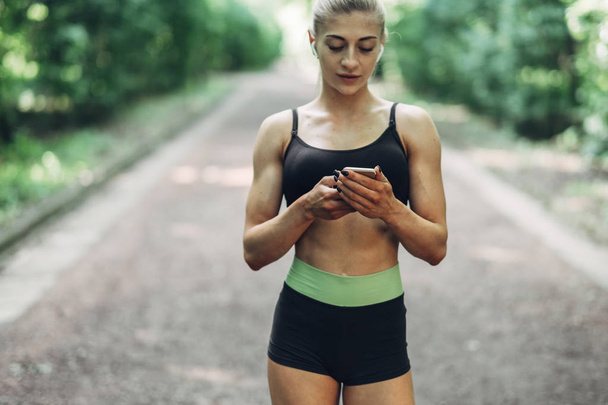 Woman Runner in the Summer Morning Park Listening to Music on Smartphone Using Bluetooth Earphones. Female Fitness Girl Jogging on Path Outside. - Photo, Image