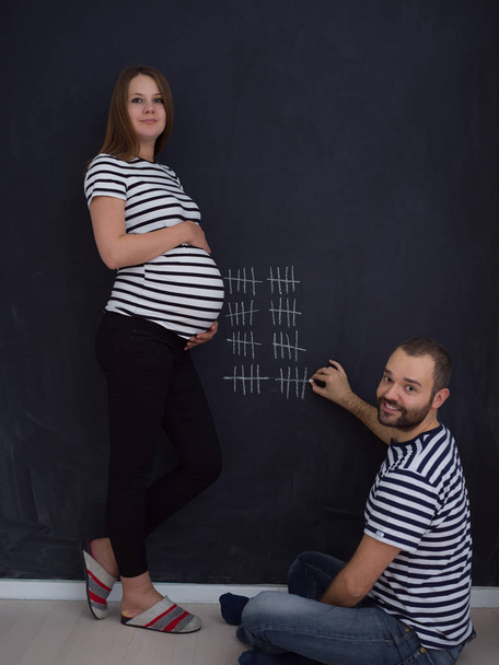 young pregnant couple accounts week of pregnancy and writing them with chalk on blackboard - Valokuva, kuva