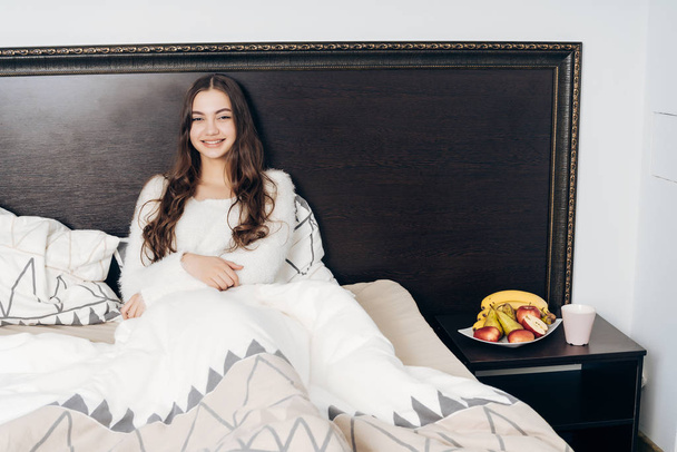 funny beautiful girl in white pajamas sits in bed, resting and wants to sleep - Fotoğraf, Görsel