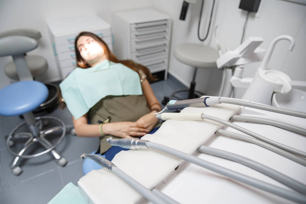 Молодая женщина сидит на стоматологическом стуле в клинике с оборудованием в мягком фокусе
. - Фото, изображение
