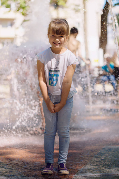 Beautiful smiling naughty little light-haired girl in jeans wearing a T-shirt walking in a sunny summer park near the fountain - Photo, image