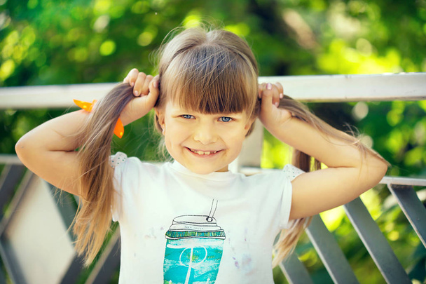 A beautiful smiling naughty little light-haired girl walks in the sunny summer park over the bridge and her hair develops in the wind - Photo, image