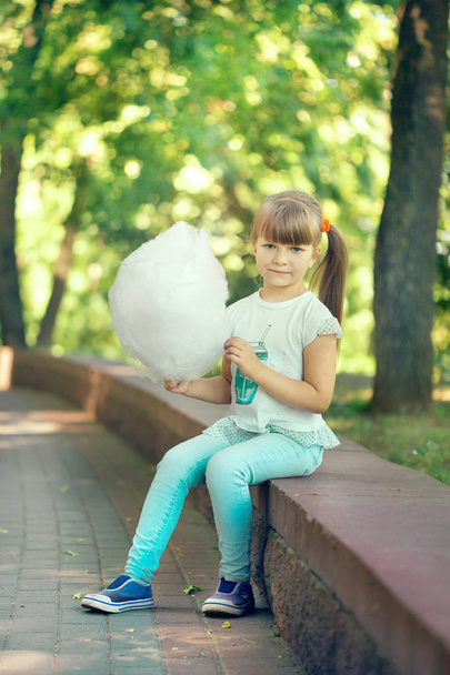 A beautiful naughty little blue-eyed light-blue girl walks in a sunny summer park and eats sweet cotton candy - Photo, image