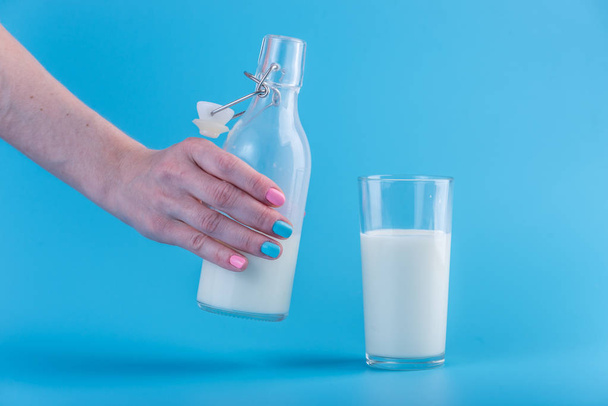Женская рука наливает свежее молоко из стеклянной бутылки в стакан на синем фоне. Красочный минимализм. Концепция здоровых молочных продуктов с кальцием
 - Фото, изображение