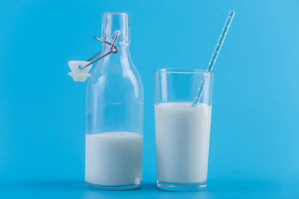 Glazen fles van verse melk en een glas met een rietje op een blauwe achtergrond. Kleurrijke minimalisme. Het concept van gezonde zuivelproducten met calcium - Foto, afbeelding