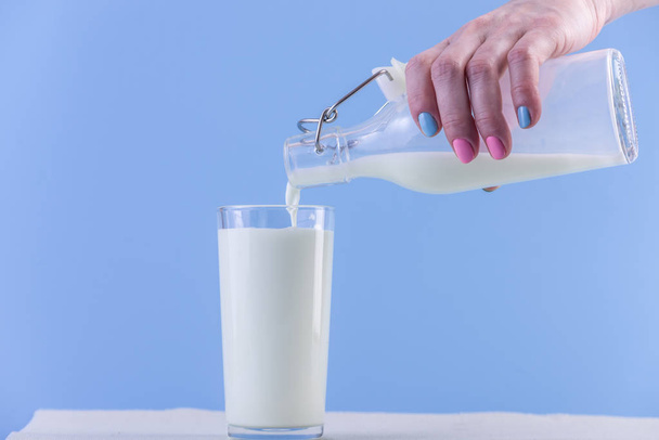 Женская рука наливает свежее молоко из стеклянной бутылки в стакан на синем фоне. Красочный минимализм. Концепция здоровых молочных продуктов с кальцием
 - Фото, изображение