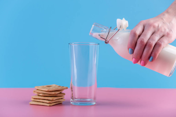 Женская рука наливает свежее молоко из стеклянной бутылки в стакан и печенье на пастельном фоне. Красочный минимализм. Концепция здоровых молочных продуктов с кальцием
 - Фото, изображение