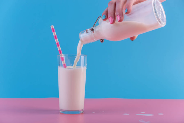 Женская рука наливает свежее молоко из стеклянной бутылки в стакан на пастельном фоне. Красочный минимализм. Концепция здоровых молочных продуктов с кальцием
 - Фото, изображение