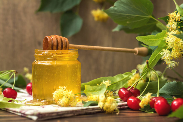 Open glass jar of liquid honey and honey dipper, bunch of linden flowers and red cherry on wooden surface. Ray of sunlight. Dark rustic style. - Photo, Image