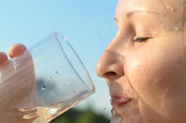 Una semplice giovane donna europea sta bevendo acqua da un bicchiere di vetro contro un cielo blu
. - Foto, immagini