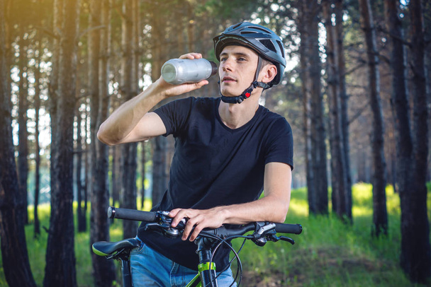 Bir bisikletçi bir kask içme suyu parkta ağaçların arasında sürme sırasında bir şişe. Etkin ve sağlıklı yaşam kavramı - Fotoğraf, Görsel