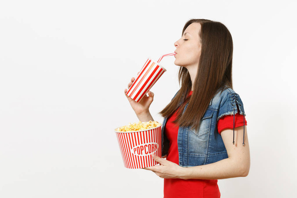 Retrato de una joven hermosa mujer con los ojos cerrados viendo películas de cine, sosteniendo un cubo de palomitas de maíz, bebiendo de una taza de plástico de refresco o cola aislada sobre fondo blanco. Emociones en el concepto de cine
 - Foto, imagen