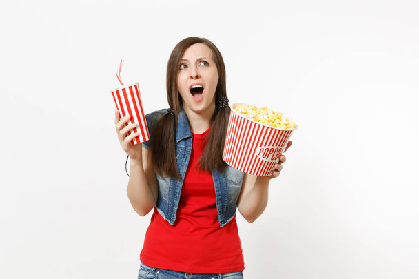 Porträt einer jungen schockierten, irritierten Frau in lässiger Kleidung, die einen Film anschaut, einen Eimer Popcorn und einen Plastikbecher Limo oder Cola in der Hand hält und isoliert auf weißem Hintergrund nach oben schaut. Emotionen im Kino - Foto, Bild