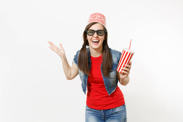 Молодая смеющаяся женщина в 3D-очках с ведром для попкорна на голове, смотрящая фильм с чашкой содовой или колой, указывающей руку в сторону на изолированном на белом фоне пространстве. Эмоции в кино
 - Фото, изображение