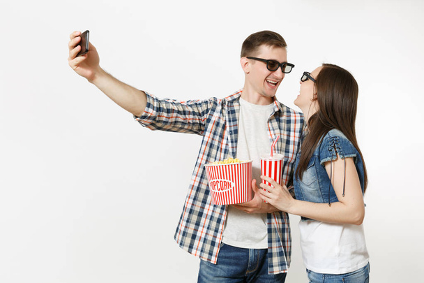 Молодая пара, женщина и мужчина в 3D очках смотрят фильм на свидании, держа ведро попкорна и чашку содовой или колы, делая селфи на мобильном телефоне, изолированном на белом фоне. Эмоции в кино
 - Фото, изображение