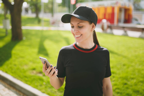 Young athletic smiling beautiful brunette girl in black uniform and cap using mobile phone during training, looking on smartphone, standing in city park outdoors. Fitness, healthy lifestyle concept - Photo, Image