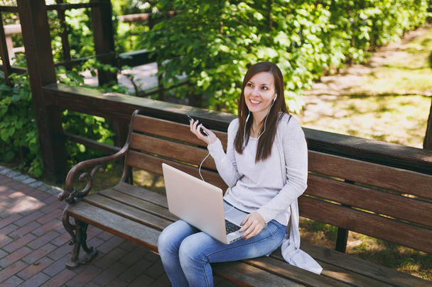 Young girl listen music in mobile phone with earphones. Woman sitting on bench working on modern laptop pc computer in city park in street outdoors on nature. Mobile Office Freelance business concept - Photo, image