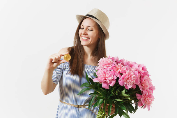 Young woman in dress, hat holding bitcoin, coin of golden color, bouquet of beautiful pink peonies flowers isolated on white background. Business, delivery, online shopping, virtual currency concept - 写真・画像