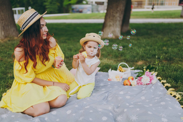 Pretty woman in yellow dress play on green grass in park, rest with little cute child baby girl hold soap bubble blower. Mother, little kid daughter. Mother's Day, love family, parenthood, childhood - Photo, Image