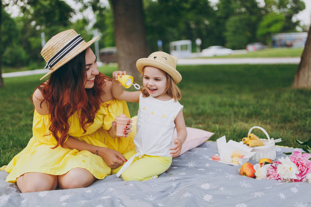 Laughing woman in yellow dress play on green grass in park rest with little cute child baby girl hold soap bubble blower. Mother, little kid daughter. Mother's Day, love family, parenthood, childhood - Photo, Image