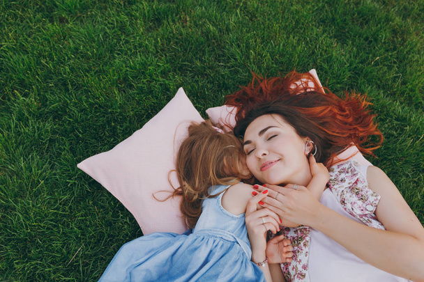 Tender woman and little cute child baby girl lie on pillows on grass in park hug, embrace and rest, have fun. Mother, little kid daughter. Mother's Day, love family, parenthood, childhood concept - Photo, Image