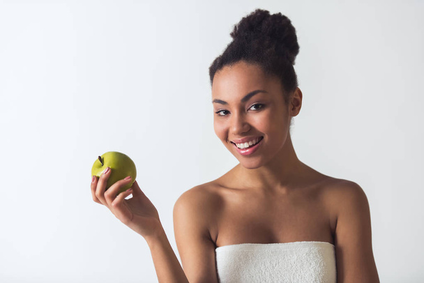 Beautiful Afro-American girl in towel is holding an apple, looking at camera and smiling, isolated on white - Photo, Image