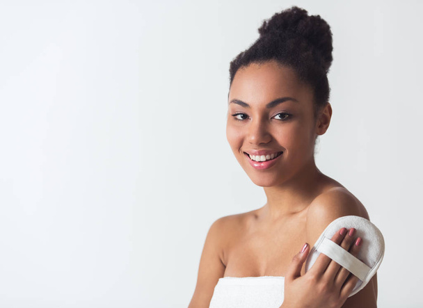 Красивая афро-американская девушка в полотенце трогает свое плечо скребком для ванны, смотрит в камеру и улыбается, изолированная на белом
 - Фото, изображение