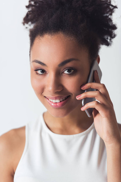 Beautiful Afro-American girl is talking on the mobile phone, looking at camera and smiling, isolated on white - Foto, Imagem