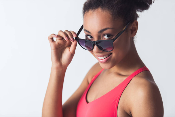 Menina afro-americana bonita em maiô rosa brilhante está olhando para a câmera de baixo dos óculos de sol e sorrindo, isolado em branco
 - Foto, Imagem