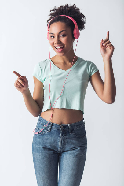 Stijlvolle Afro-Amerikaanse meisje in casual kleding en hoofdtelefoon is luisteren naar muziek, kijken naar camera en glimlachend, geïsoleerd op wit - Foto, afbeelding