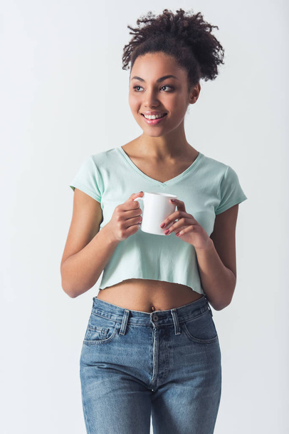 Cheerful Afro-American girl in casual clothes is holding a cup, looking away and smiling, isolated on white - 写真・画像