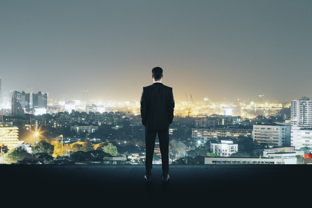 Visão traseira do jovem empresário no telhado olhando para a cidade noturna iluminada. Conceito de investigação e procura de emprego
  - Foto, Imagem