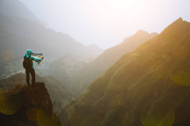 Człowiek z mapą pozostający na szczycie górskiej skały z pięknym widokiem na wysokie pasma górskie i głęboką dolinę - Zdjęcie, obraz