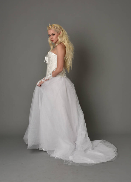 full length portrait of blonde  girl wearing white fantasy gown. standing pose in side profile, grey studio background. - Photo, Image