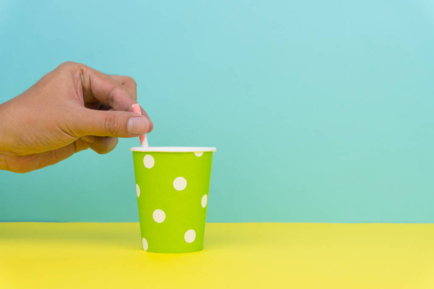 put a pink striped straw into a green polka dot party paper cup, yellow table with blue background - Photo, image