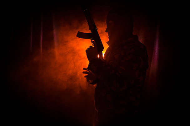 Σιλουέτα του ανθρώπου με την επίθεση τουφέκι έτοιμος να επιτεθεί σε σκούρο φόντο τονισμένο ομίχλη ή επικίνδυνο ληστή σε μαύρο, φορώντας κουκούλα και εκμετάλλευση όπλο στο χέρι. Γυρίσματα τρομοκράτης με το όπλο θέμα ντεκόρ - Φωτογραφία, εικόνα