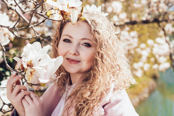 luxurious curly woman posing in a park in the sun next to a blooming magnolia, smiling - Фото, зображення