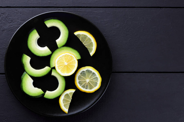 Avocado on a black plate top view, sliced avocado and lemon on black wooden boards, vegetarian food, useful natural food, tropical fruits for breakfast, copy space - Photo, image