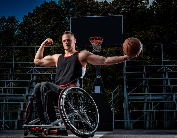 Fuerte jugador de baloncesto en silla de ruedas posar con una pelota en campo de juego abierto
. - Foto, Imagen