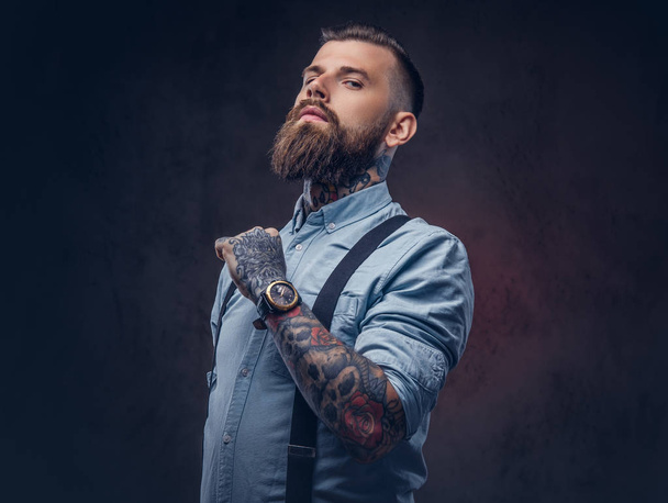 Πορτρέτο του ένα όμορφος ντεμοντέ hipster σε ένα μπλε πουκάμισο και ζαρτιέρες. Απομονωμένα σε σκούρο φόντο. - Φωτογραφία, εικόνα