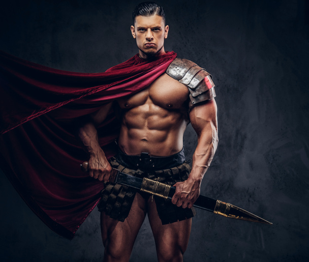 Brutal ancient Greek warrior with a muscular body in battle equipment posing on a dark background. - Фото, изображение