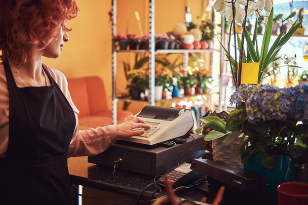 Charmoso ruiva florista feminina vestindo uniforme trabalha com uma caixa registadora na loja de flores
. - Foto, Imagem