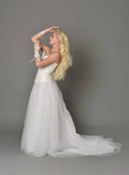 full length portrait of blonde  girl wearing white fantasy gown. standing pose in side profile, grey studio background. - Foto, imagen