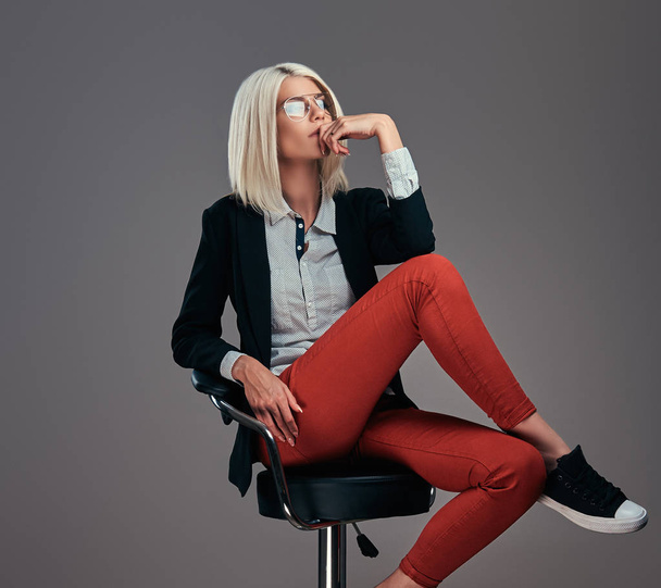 Mode blonde vrouw in trendy kleding en bril poseren in de studio zittend op een stoel. Geïsoleerd op een grijze achtergrond. - Foto, afbeelding