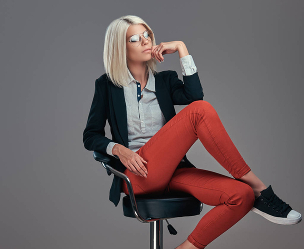 Mulher loira moda em roupas da moda e óculos posando no estúdio enquanto se senta em uma cadeira. Isolado em um fundo cinza
. - Foto, Imagem