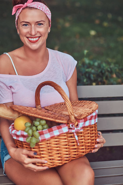 Szczęśliwy piękne rude kobiet na sobie ubranie posiada kosz piknikowy siedząc na ławce w parku. - Zdjęcie, obraz